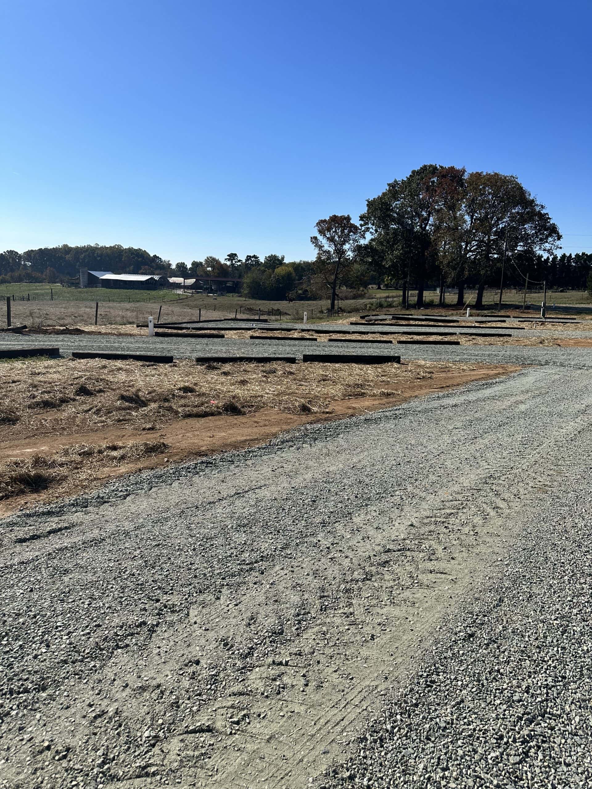 A gravel road leads through a rural landscape under a clear blue sky. On the left, wooden beams lay scattered in a field. In the background, trees with autumn leaves stand near a barn. A wire fence runs alongside the road.