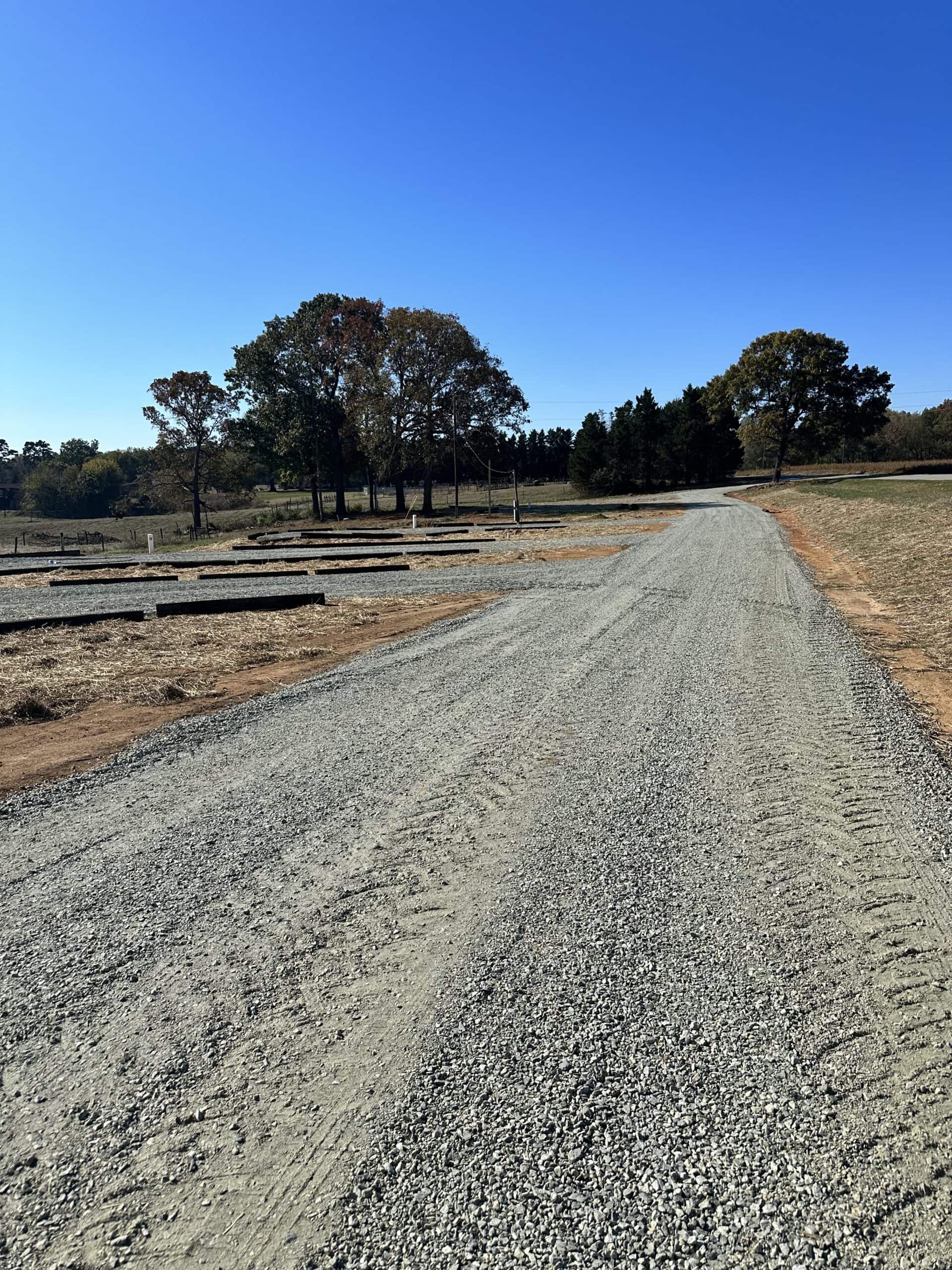 A gravel road runs through a rural area, surrounded by dry grass and trees under a clear blue sky. Construction materials are scattered on the left side, suggesting ongoing development or maintenance work.